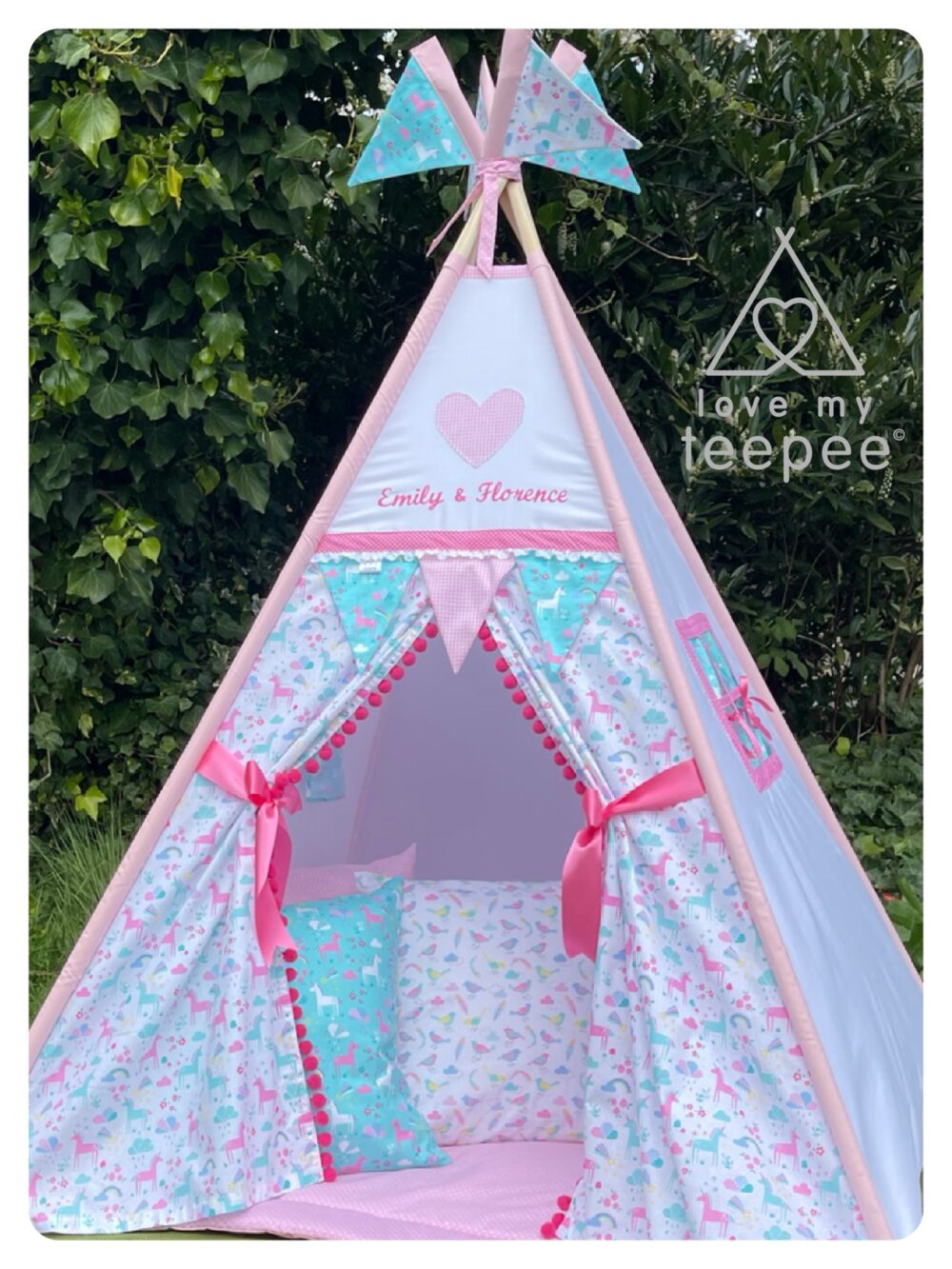 Handmade Kids Teepee Tent Unicorn Forest Pink & Aqua. White teepee with doors patterned with unicorns, rainbows, and clouds in pinks, aqua, and turquoise, trimmed with pink pom-poms. Above the doors is a heart motif with dark pink trim, optional embroidered names above the doors (example: Emily and Florence), and four decorative flags. Includes ribbon tie-backs, matching scatter cushions, and padded floor mat.