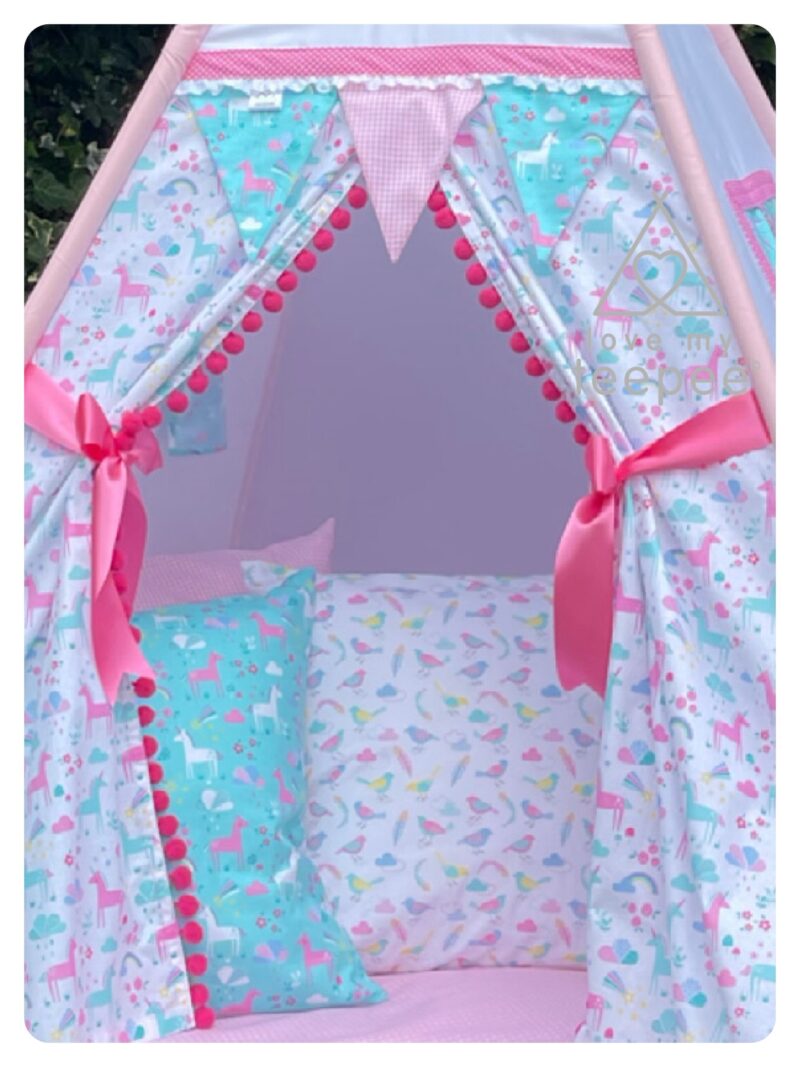 Close-up of Unicorn Forest Pink & Aqua teepee doors patterned with unicorns, rainbows, and clouds in pinks, aqua, and turquoise. Shows pom-pom edging, ribbon tie-backs, matching scatter cushions, padded floor mat, and optional embroidered names above the doors (example: Emily and Florence).