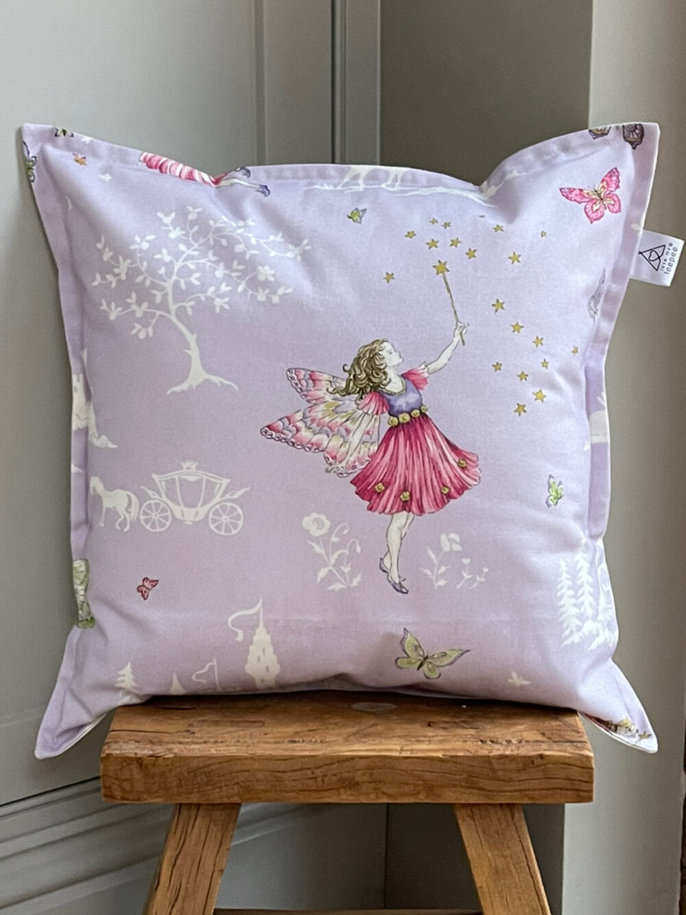 Handmade Fairyland cushion in lilac by Sanderson, featuring delicate fairy, floral and butterfly fabric, with optional pom-pom edging, styled for a cosy children’s space